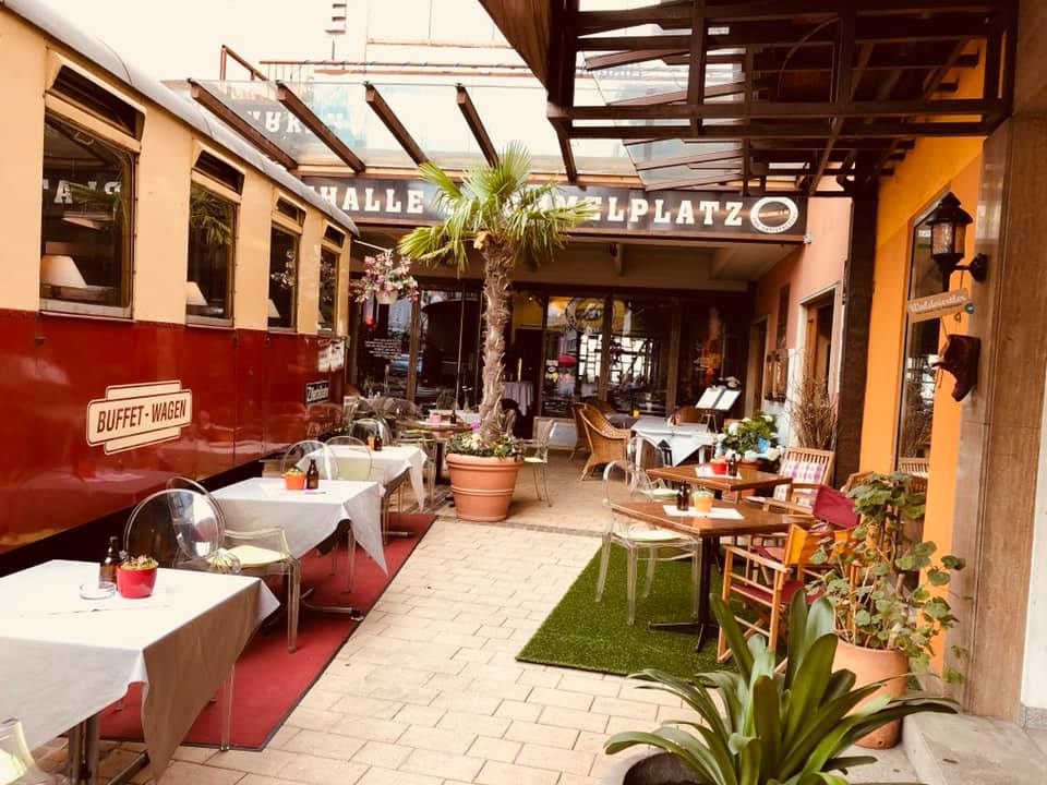 Restaurantterrasse mit Eisenbahnwagen, gedeckte Tische, Palmen, Sitzbereich im Innenhof.